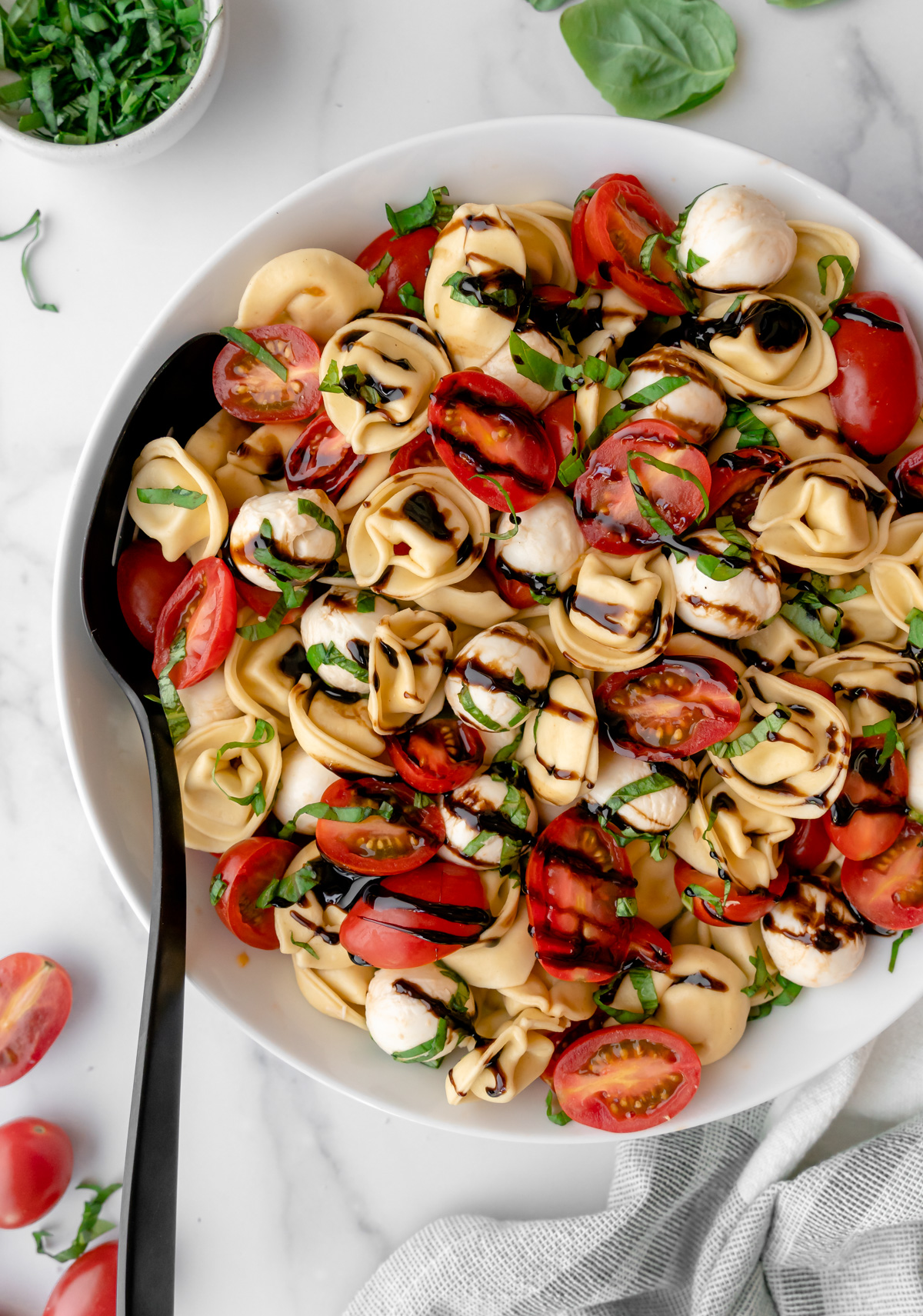Bowl of Tortellini Caprese salad with black serving spoon and basil and tomatoes on the side.