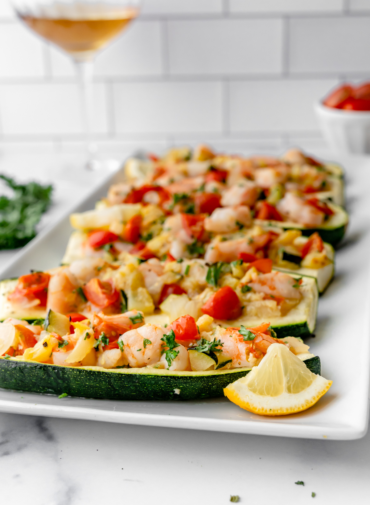 Angled view of shrimp stuffed zucchini boats with a lemon wedge and glass of wine in the back.