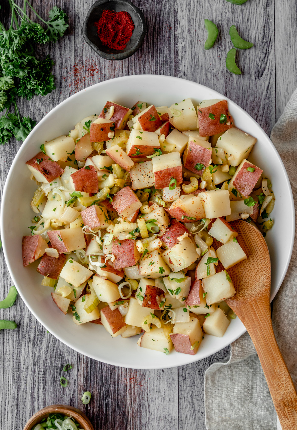 Bowl of german potato salad with a wooden serving spoon and ingredients to the side.
