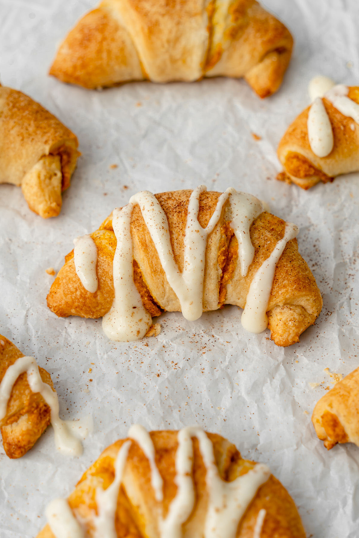 Close up on pumpkin cream cheese crescent rolls on parchment paper and drizzled with cream cheese frosting.