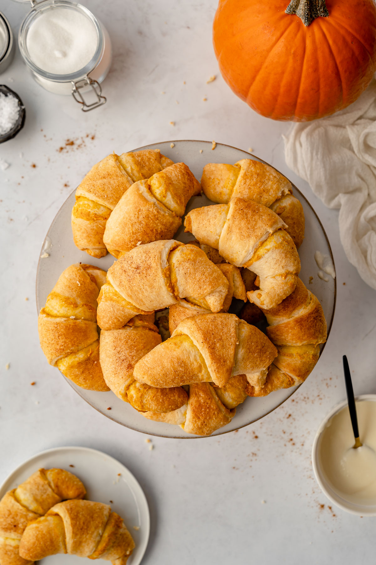 Pumpkin cream cheese crescent rolls on a cake stand with frosting, plates, pumpkin, and linen on the side.