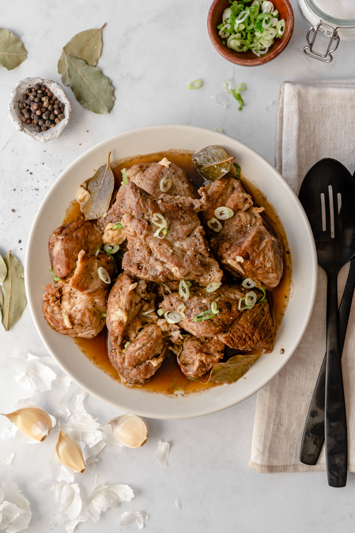 A serving bowl of Instant Pot Pork Adobo with serving utensils, garlic cloves, and bay leaves to the side.
