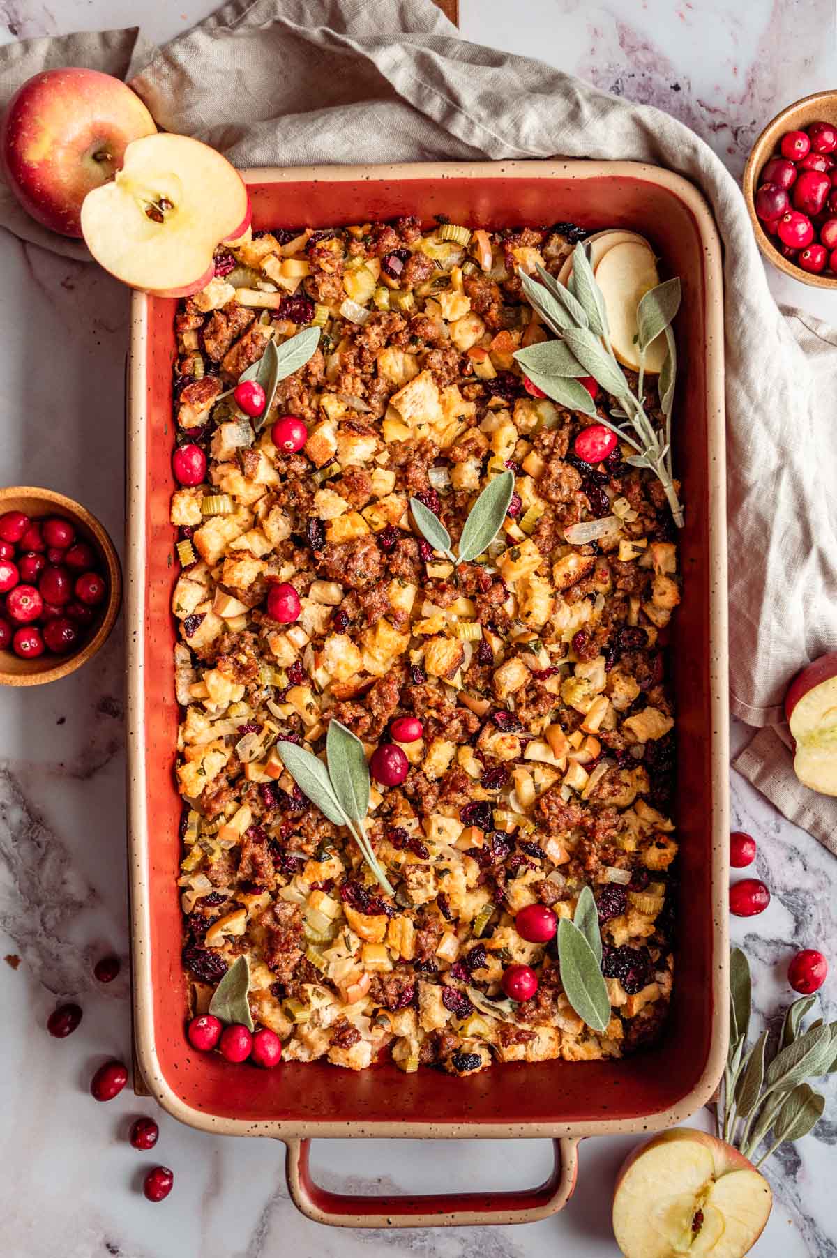 Sausage Apple Cranberry Stuffing in a red baking dish surrounded by fresh cranberries, apples, and sage leaves.