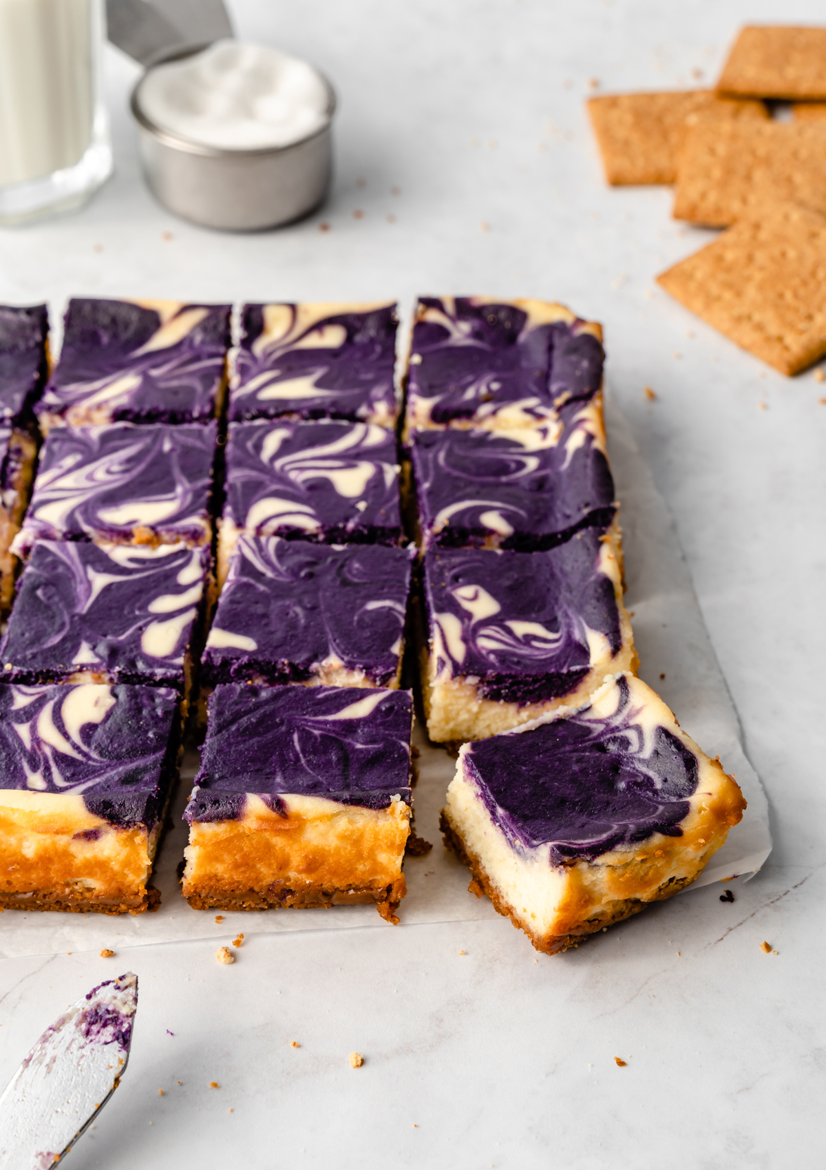 Ube cheesecake bars on parchment paper with glass of milk, cup of sugar, and knife.