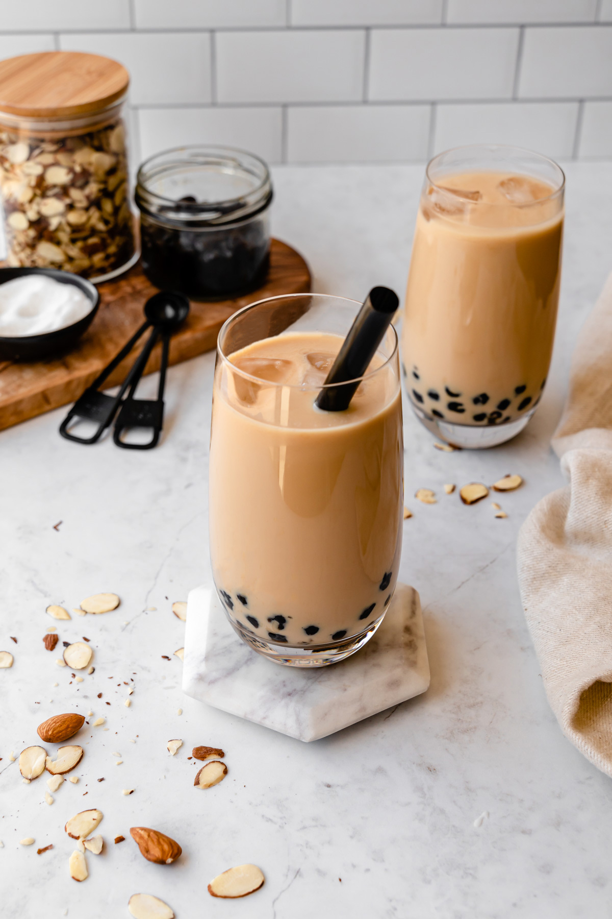 Two glasses of almond milk tea with almonds, boba, and sugar on the side.