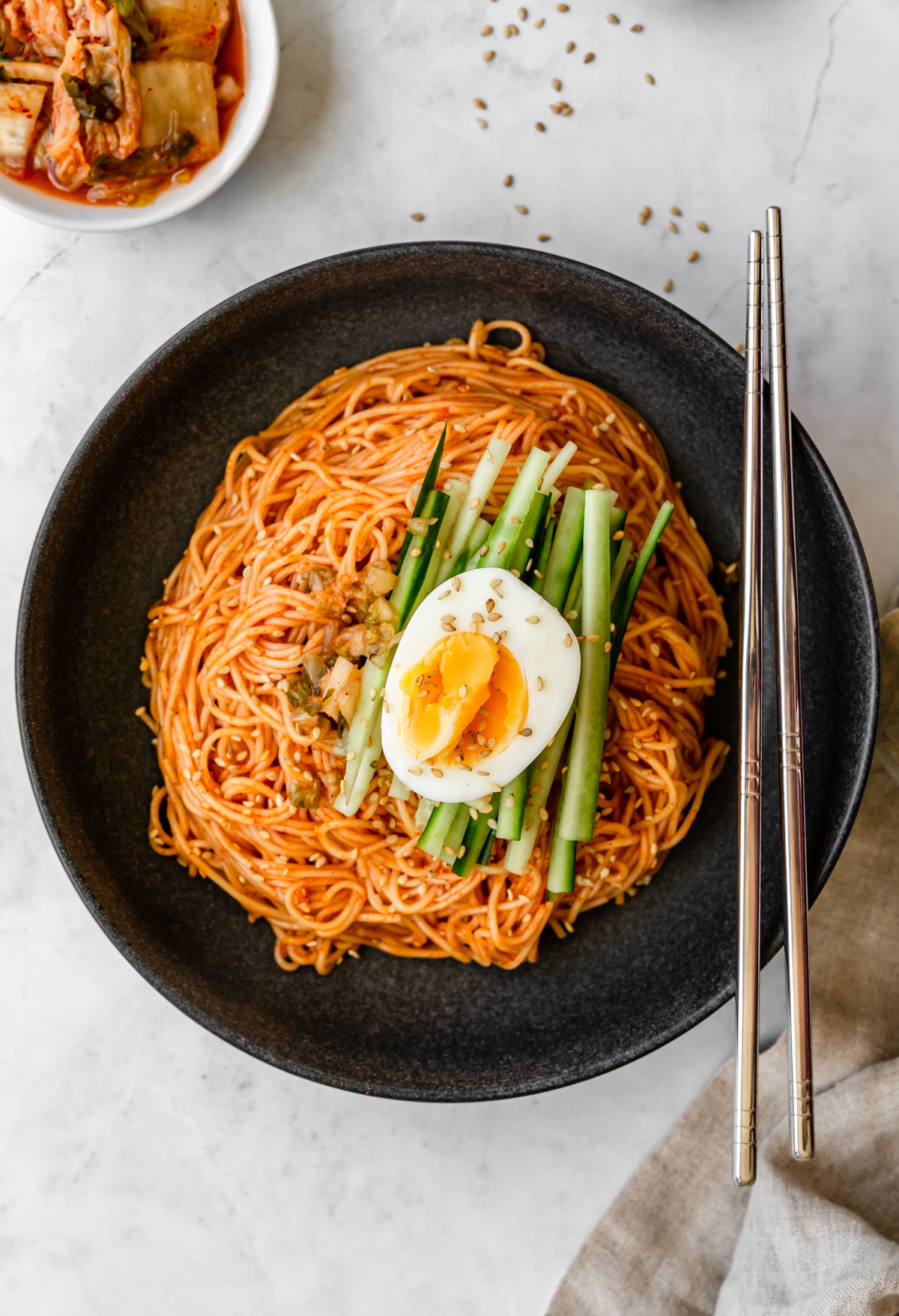 Black bowl of Korean spicy noodles topped with kimchi, cucumber, and hard boiled egg with metal chopsticks.