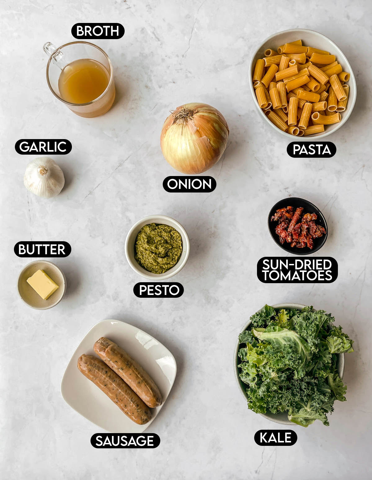 Labeled ingredients needed for sausage kale pasta: broth, garlic, onion, pasta, butter, pesto, sun-dried tomatoes, sausage, and kale.