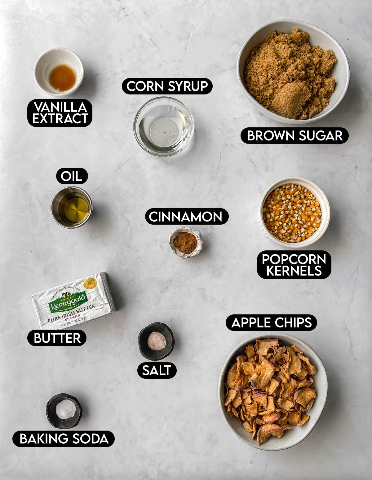 Labeled ingredients for caramel apple popcorn: vanilla extract, corn syrup, brown sugar, oil, cinnamon, popcorn kernels, butter, salt, apple chips, and baking soda.