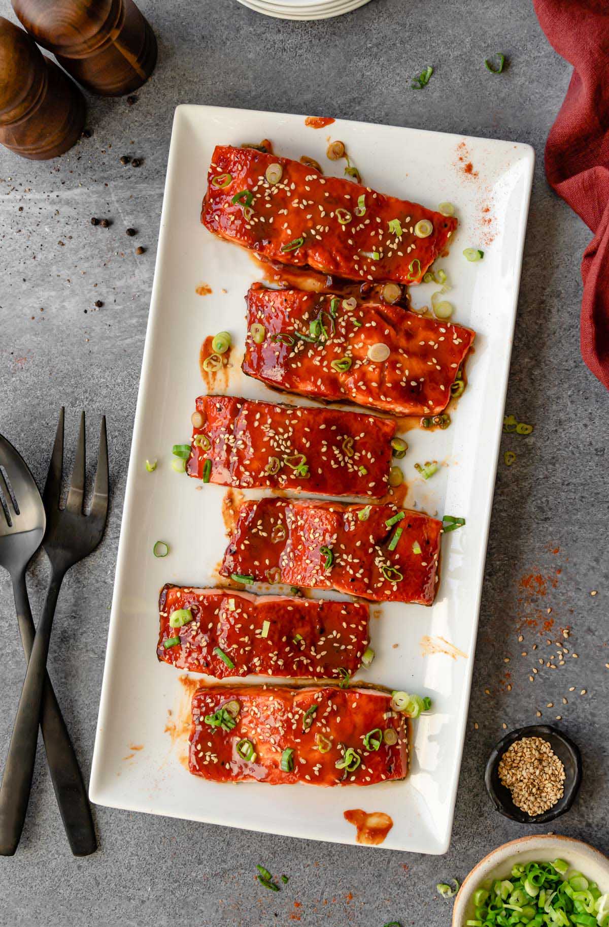 6 Gochujang Salmon fillets on a white platter with serving utensils, green onion, and plates on the side.