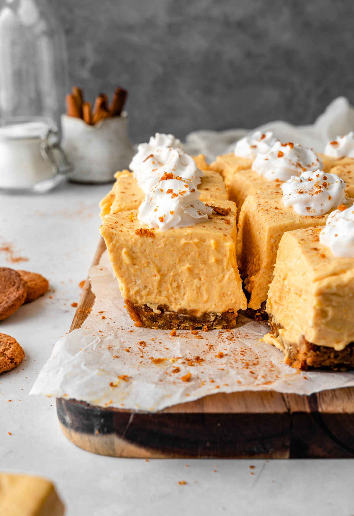 no bake pumpkin cheesecake bars on a wooden board with cookies and cinnamon sticks in the back.