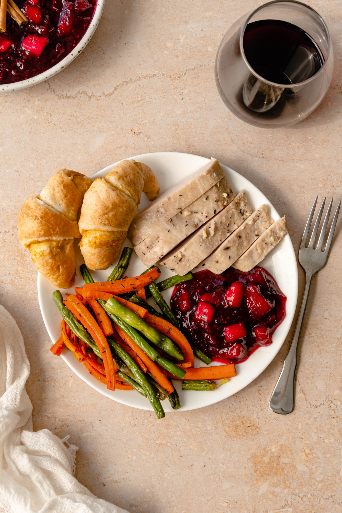 A plate of pumpkin rolls, turkey, roasted carrots and green beans, and cranberry sauce with wine and a fork on the side.