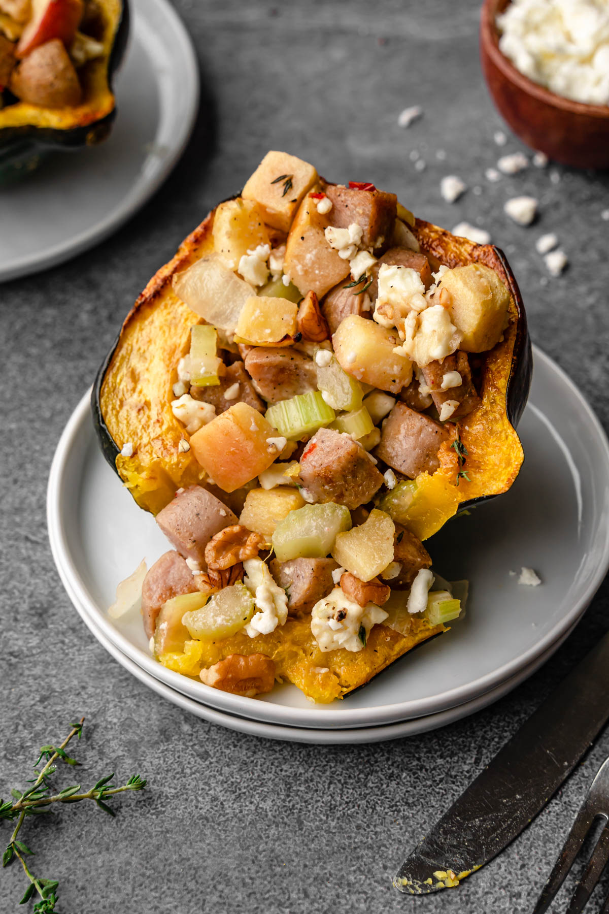 Stuffed acorn squash on a plate with a fnife, thyme, and a bowl of feta cheese beside it.