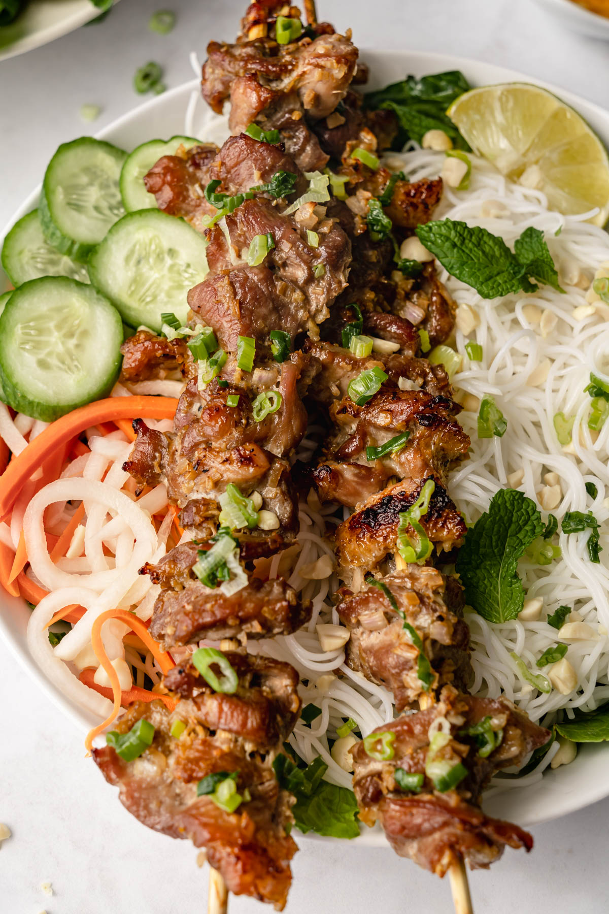 Close up of pork skewers on a bowl with rice noodles, vegetables, lime, and rice noodles.