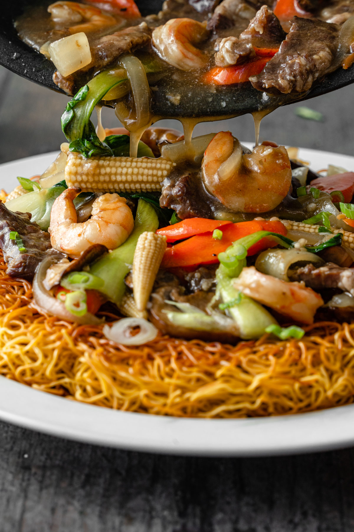 Pouring gravy with meat and vegetables on top of crispy noodles on a plate.