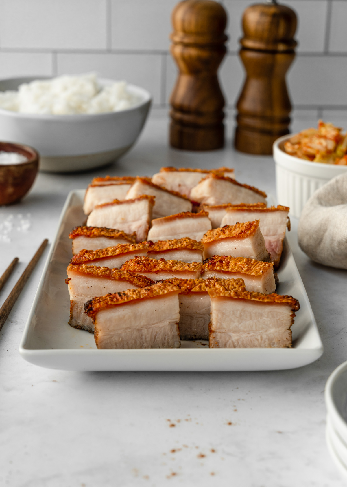 Thin slices of Vietnamese Crispy Pork Belly on a platter with rice, kimchi, chopsticks, salt, and pepper, and a napkin around it.