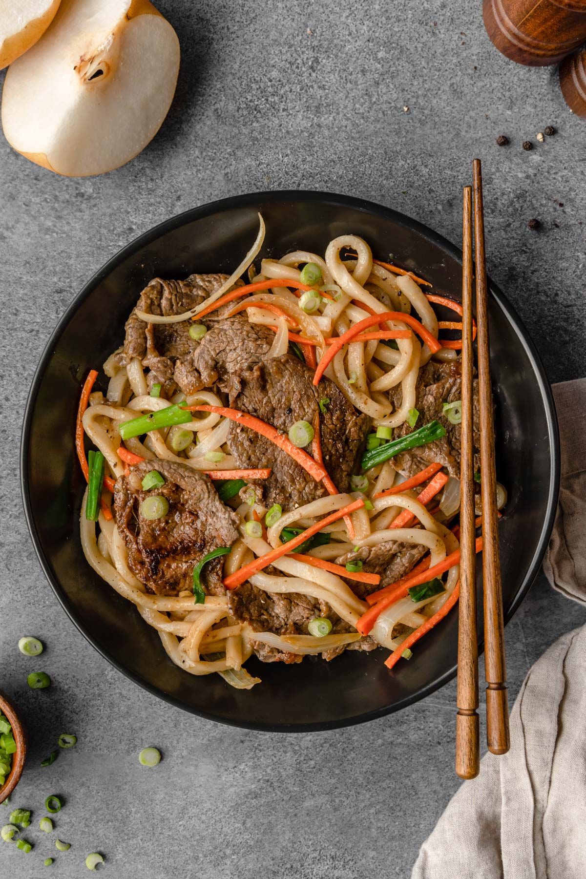 A bowl of Bulgogi Udon stir fry with chopsticks on it, and green onion and pear on the side.