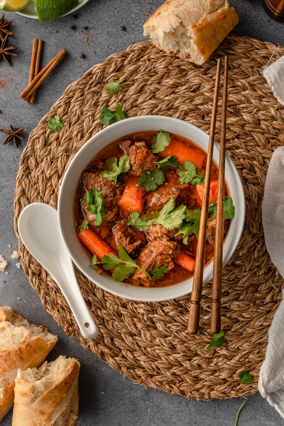 A bowl of Instant Pot Bo kho with wooden chopsticks on a brown placemat and surrounded by bread, spices, and a linen.