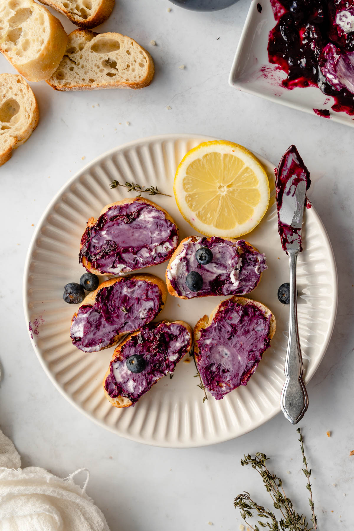 A plate of baguette slices swith Blueberry Goat Cheese spread on top with a lemon slice and butter knife to the side.