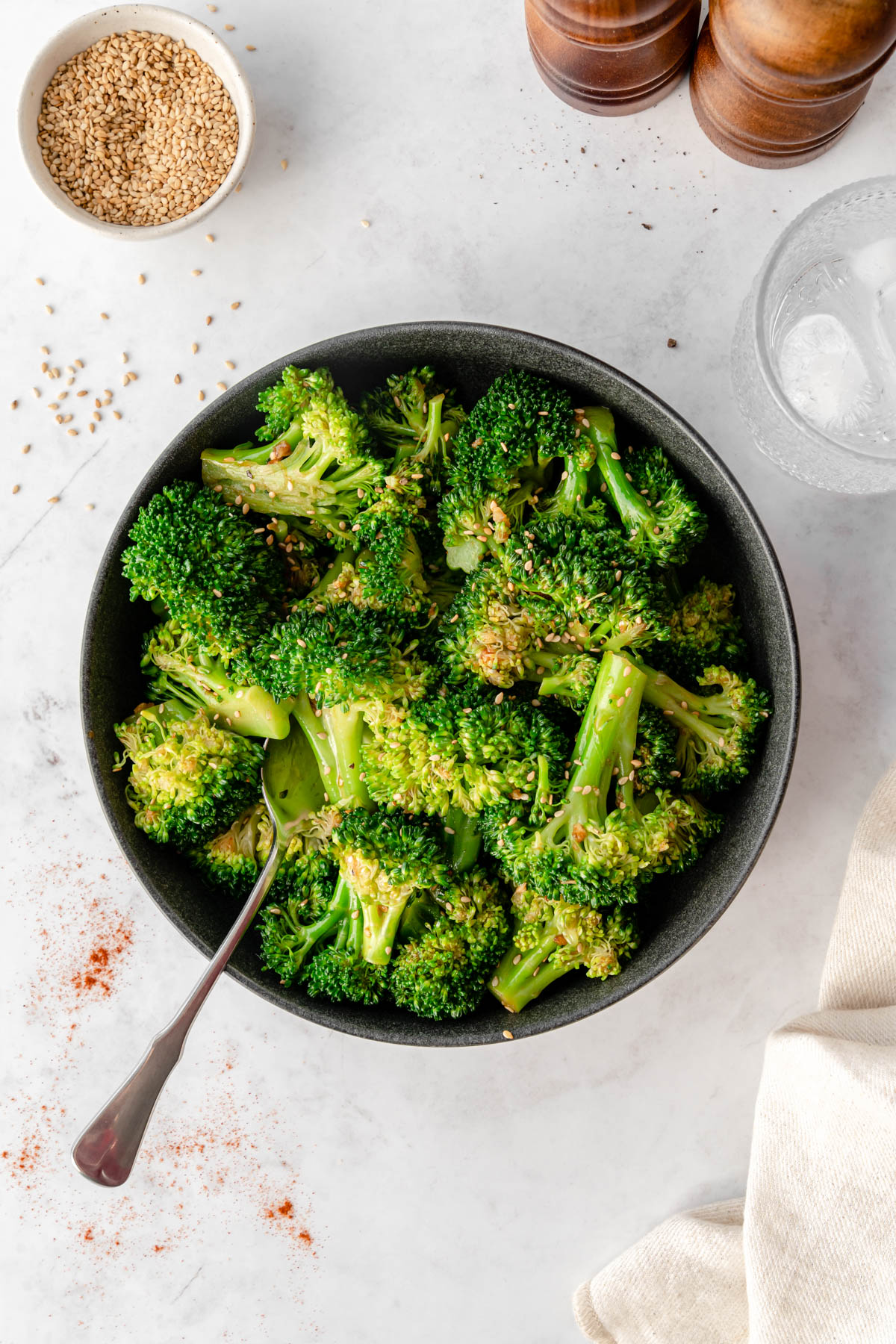 A black bowl of Korean Broccoli Salad with a spoon in it, surrounded by sesame seeds, gochugaru, a linen, ice water, and salt and pepper grinders.