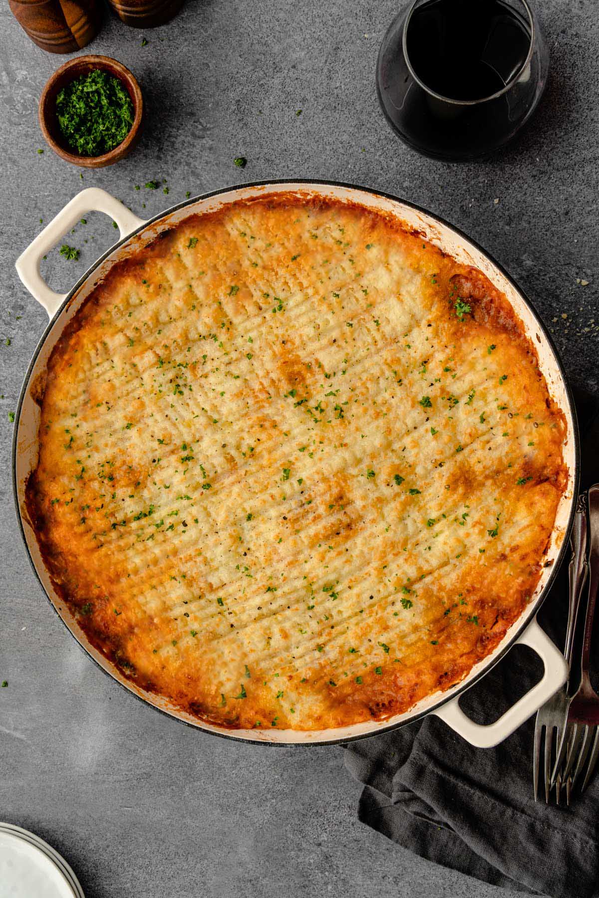 A large casserole dish of Shepherd's Pie with forks, red wine, and chopped parsley on the side.