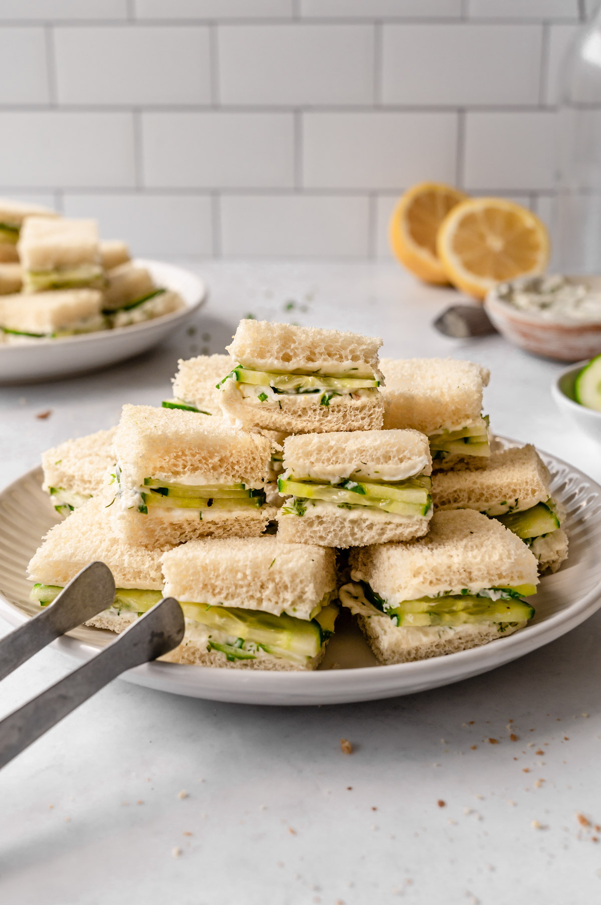 Two plates of cucumber sandwiches squares with tongs.